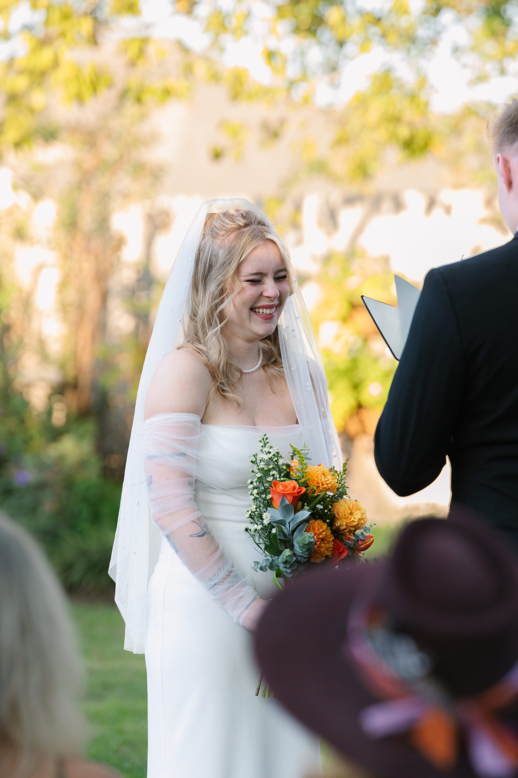 Smiling bride holding a bouquet of orange and yellow flowers during an outdoor ceremony